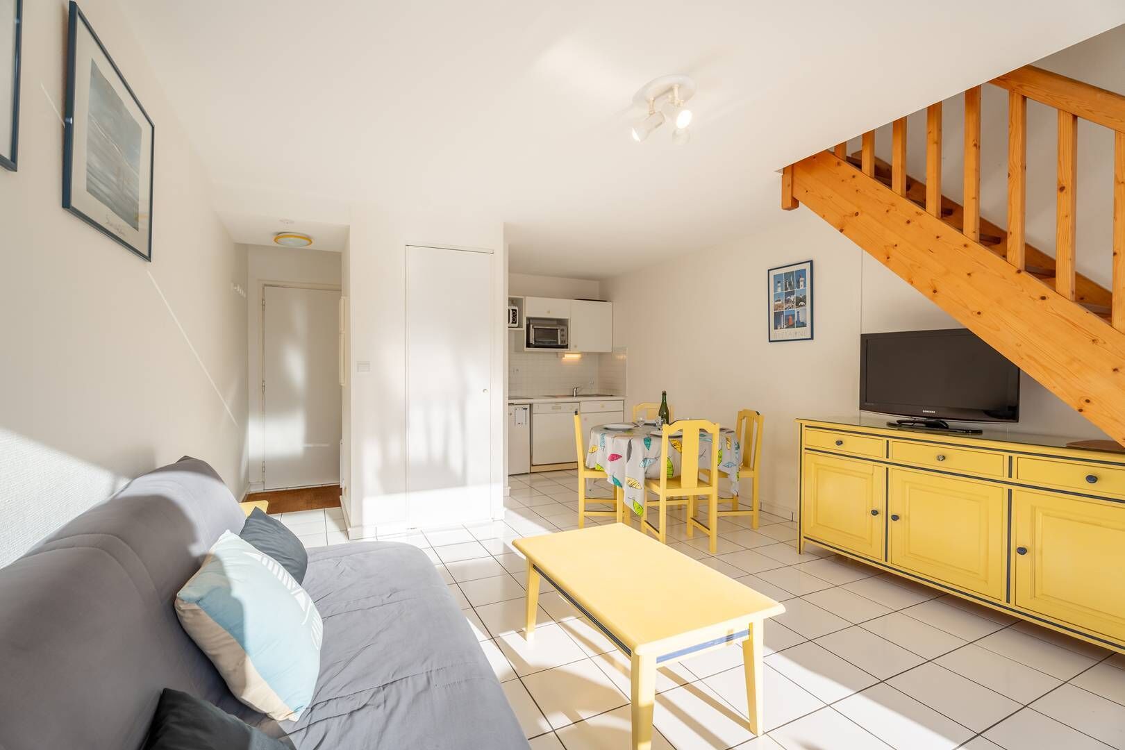 Una sala de estar acogedora con un sofá, una mesa amarilla y un televisor sobre un mueble amarillo. Se ve una escalera que sube a otro nivel.