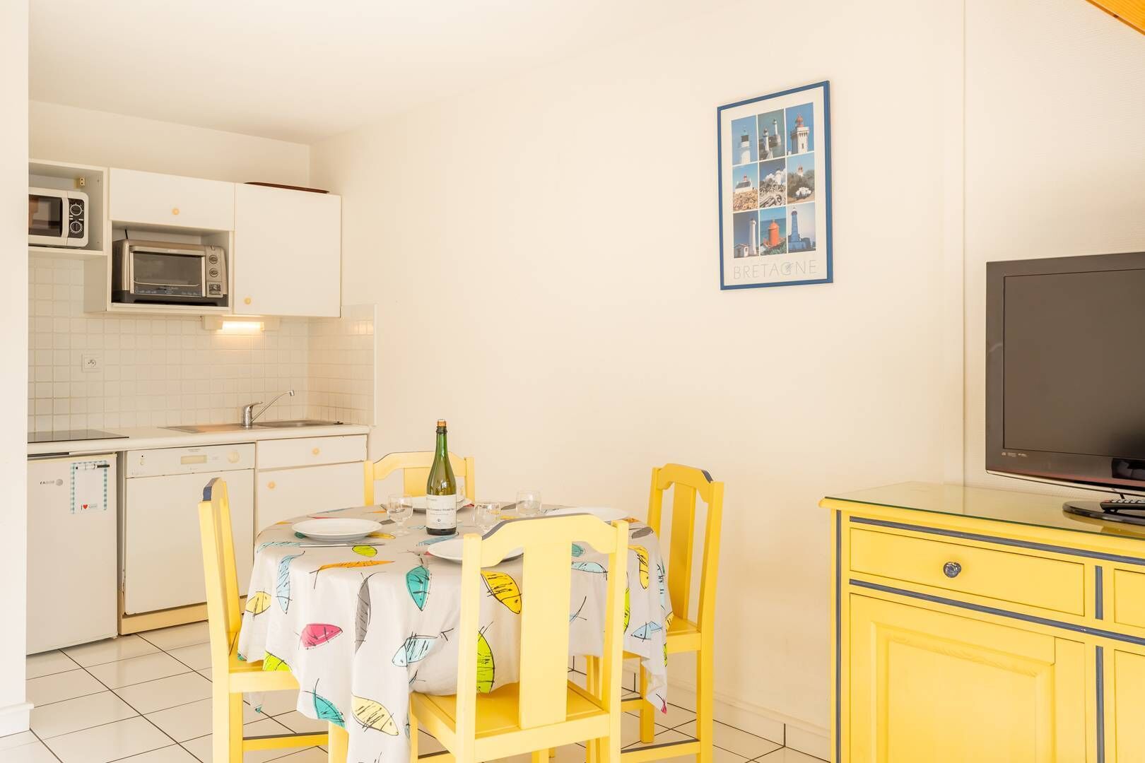 A compact kitchen with white cabinets, a microwave, and a built-in oven. A dining table with a colorful tablecloth and two chairs is set for two. A bottle of wine and glasses are on the table. A framed picture of lighthouses hangs on the wall.