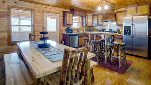 A wooden cabin interior with a kitchen area featuring a stainless steel refrigerator, wooden bar stools, and a dining table with a placemat.