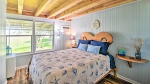 Een slaapkamer met een houten plafond, een bed met een beddeken met een patroon, een nachtkastje met een lamp en een raam met uitzicht op groen.