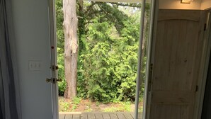 A bedroom with a wooden floor, a bed with a white cover, a door leading to an outdoor area with a wooden deck and greenery. - Spring and Summer are on the way! Book your stay Today! (West Cape May)