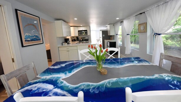 A dining area with a table covered in a wave-themed tablecloth, white chairs, a vase with red tulips, and a kitchen in the background. - Spring and Summer are on the way! Book your stay Today! (West Cape May)