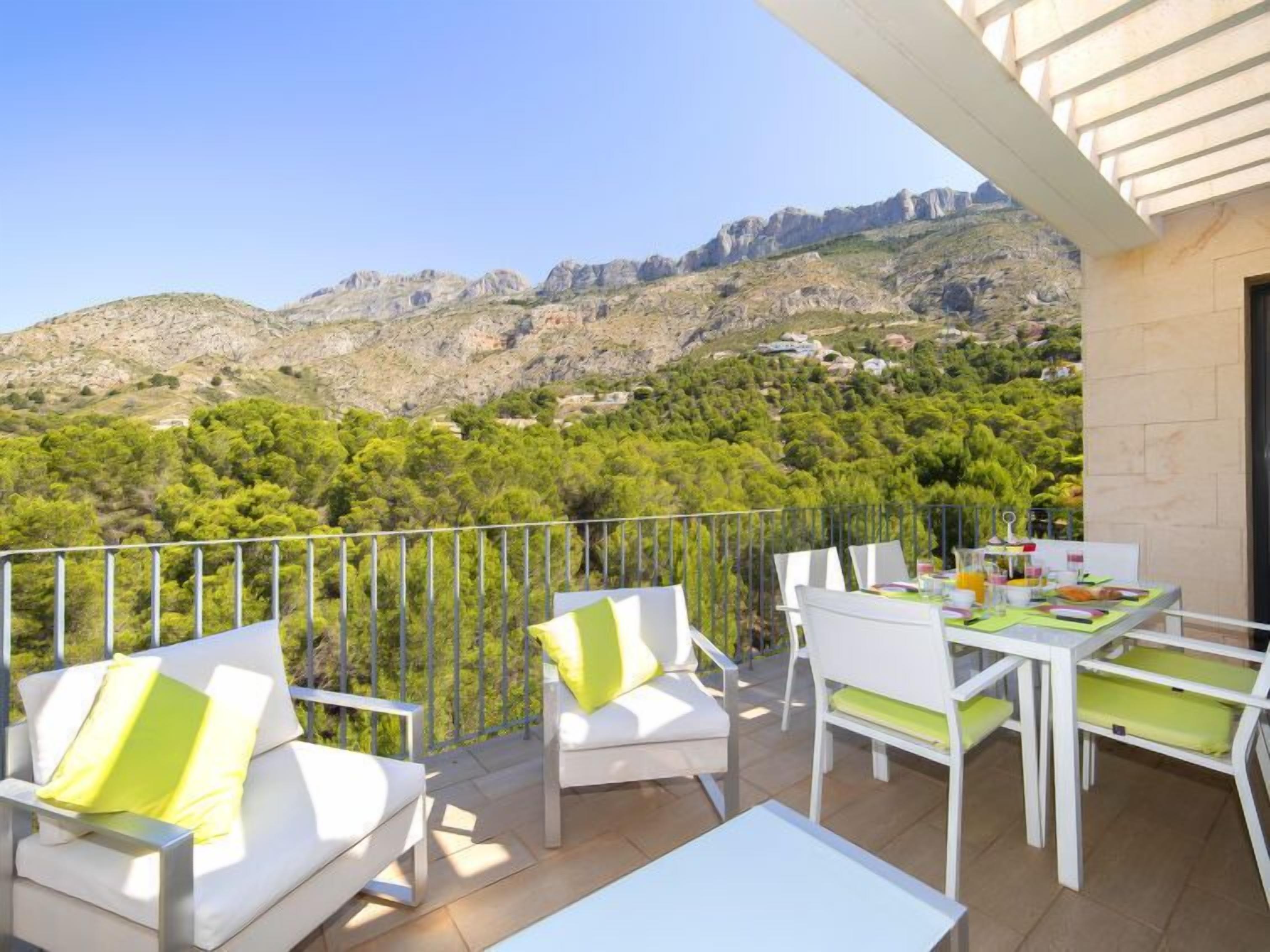 A balcony with white furniture, a table set for breakfast, and a scenic view of mountains and greenery.