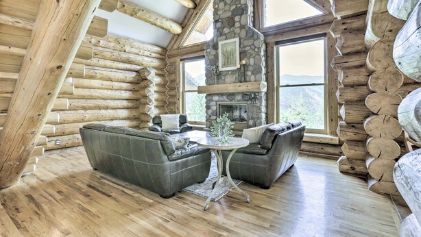 A log cabin interior with a stone fireplace, leather sofas, and large windows offering a mountain view. - Mountain Retreat w/ Deck: Ski, Soak, Golf & Hike! (Glenwood Springs)