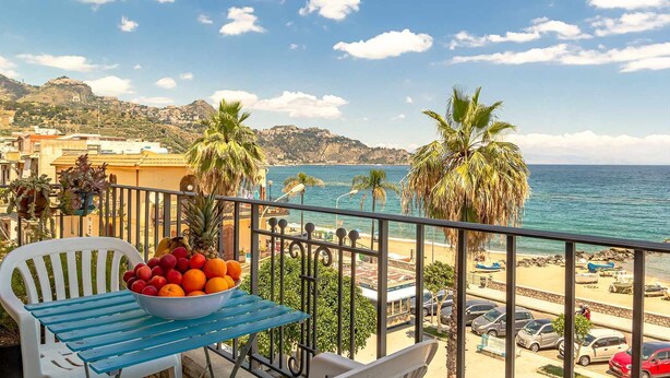 A balcony with a table, a bowl of fruit, and a view of the sea and beach. - La Casa di Elvira - Sea View (Giardini Naxos)