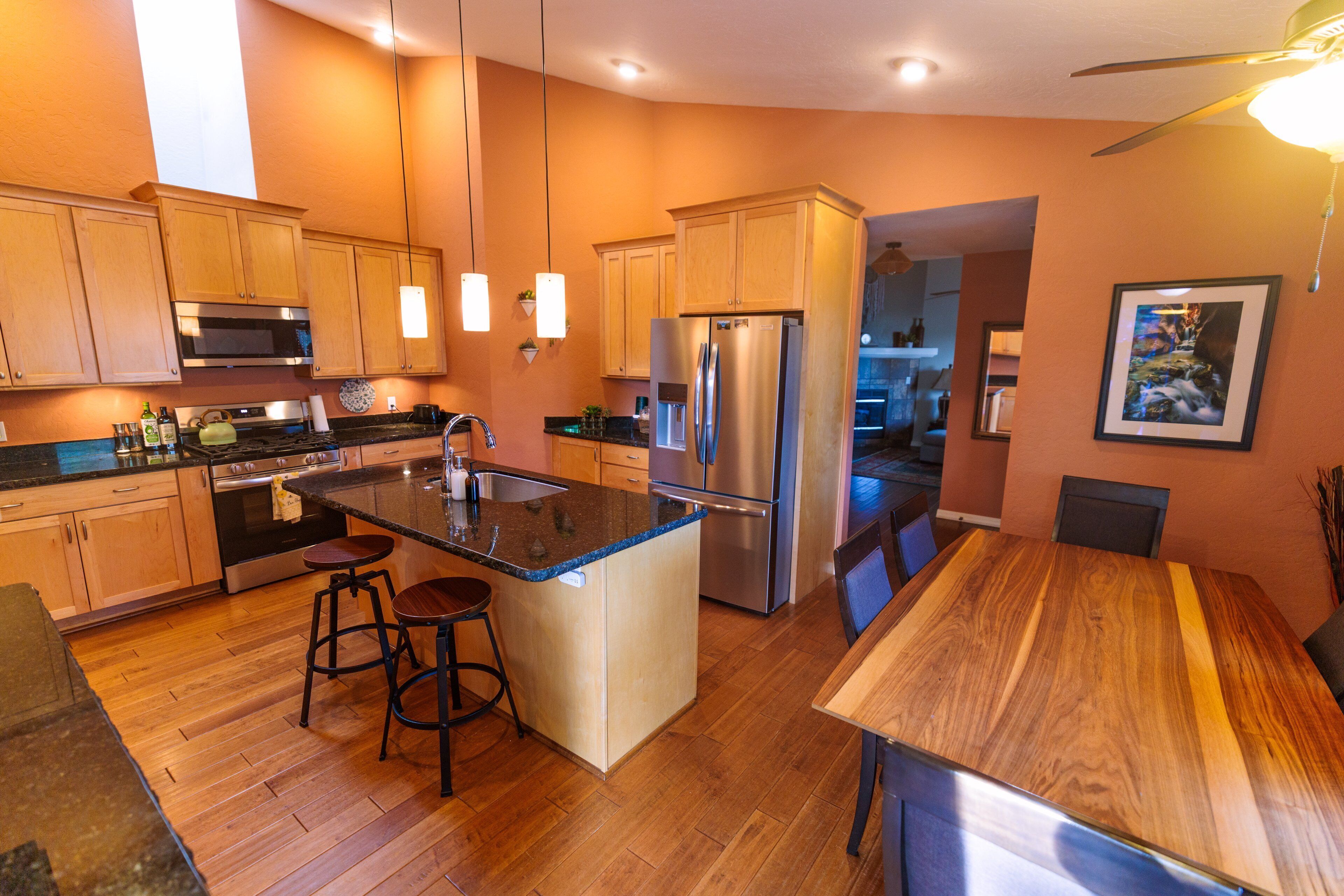 A modern kitchen with wooden cabinets, stainless steel appliances, a central island with stools, and a dining area with a wooden table.