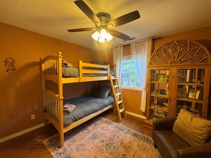 A room with a bunk bed, a ceiling fan, a window with curtains, a patterned rug, and a wooden cabinet with glass doors.