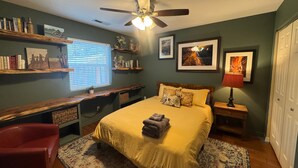 A bedroom with a bed, a desk, shelves with books, a ceiling fan, and artwork on the walls.