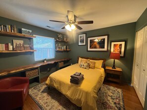 A bedroom with a bed, a desk, shelves with books, a ceiling fan, and artwork on the walls.