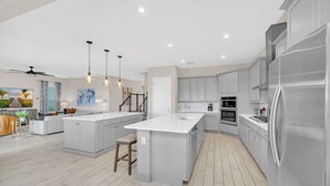 A modern kitchen with grey cabinetry, a large island, stainless steel appliances, pendant lighting, and a view of a living area with a sofa and plants. - Paradise at Bear's Den | 6-Bed Villa w/ Loft, Games Room, and Private Pool (Kissimmee)