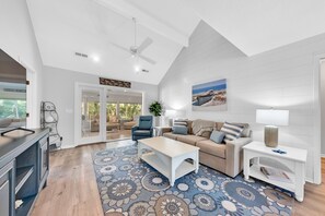 Un salon spacieux avec un plafond haut, un ventilateur de plafond, une grande fenêtre, un ensemble de canapés, une table basse, une table d’appoint et un tapis à motifs.