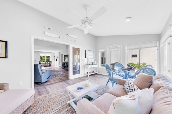 A spacious living area with a sofa, coffee table, and blue chairs. There is a ceiling fan and a door leading to another room.