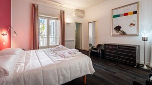 Une chambre d’hôtel avec un grand lit, un bureau et une chaise. Il y a une fenêtre avec des rideaux, un radiateur et un tableau au mur.