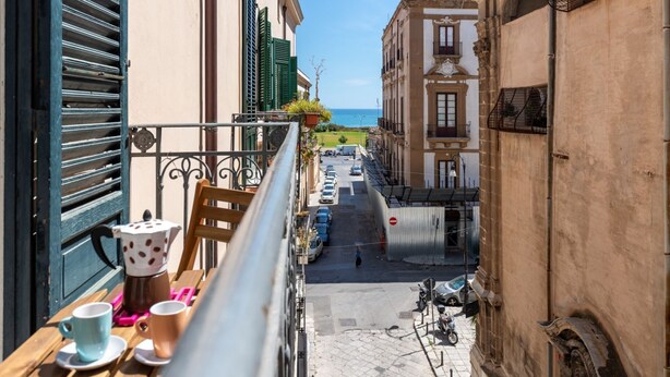 Une rue étroite bordée de bâtiments des deux côtés, une table en bois avec des tasses et une cafetière, et une vue sur la mer au loin.