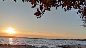 Ein Strand bei Sonnenuntergang, im Vordergrund üppiges Grün und rosafarbene Blüten, das Meer liegt ruhig.