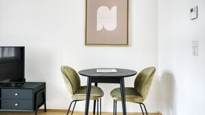 Een kamer met een zwarte tv-standaard, een klein tafeltje met twee stoelen en een ingelijst kunstwerk aan de muur.