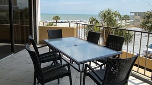 Een balkon met een glazen tafel en stoelen, met uitzicht op het strand en de oceaan.