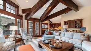 Un salon spacieux avec un grand canapé, une table basse et une cheminée. Le plafond est orné de poutres en bois et de grandes fenêtres.