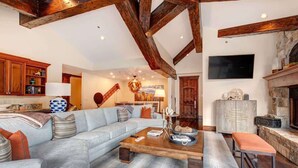 Un salon spacieux avec un grand canapé, des poutres en bois, une cheminée et un téléviseur fixé au mur.