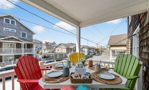 A balcony with a table set for dining, featuring Adirondack chairs, a sign, and a view of neighboring houses.