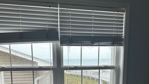 Un sofá gris frente a una ventana con persianas, con vista a una terraza de madera y al océano.