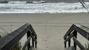 Una escalera de madera que lleva a una playa arenosa con olas oceánicas agitadas al fondo.