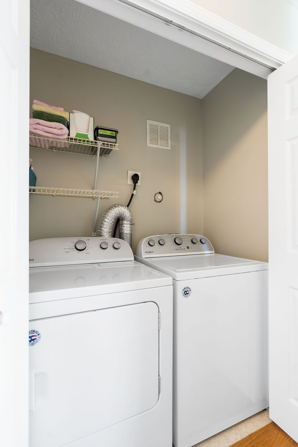 A laundry room with two front-loading washers and dryers, a metal shelf with folded towels, and a vent on the wall.