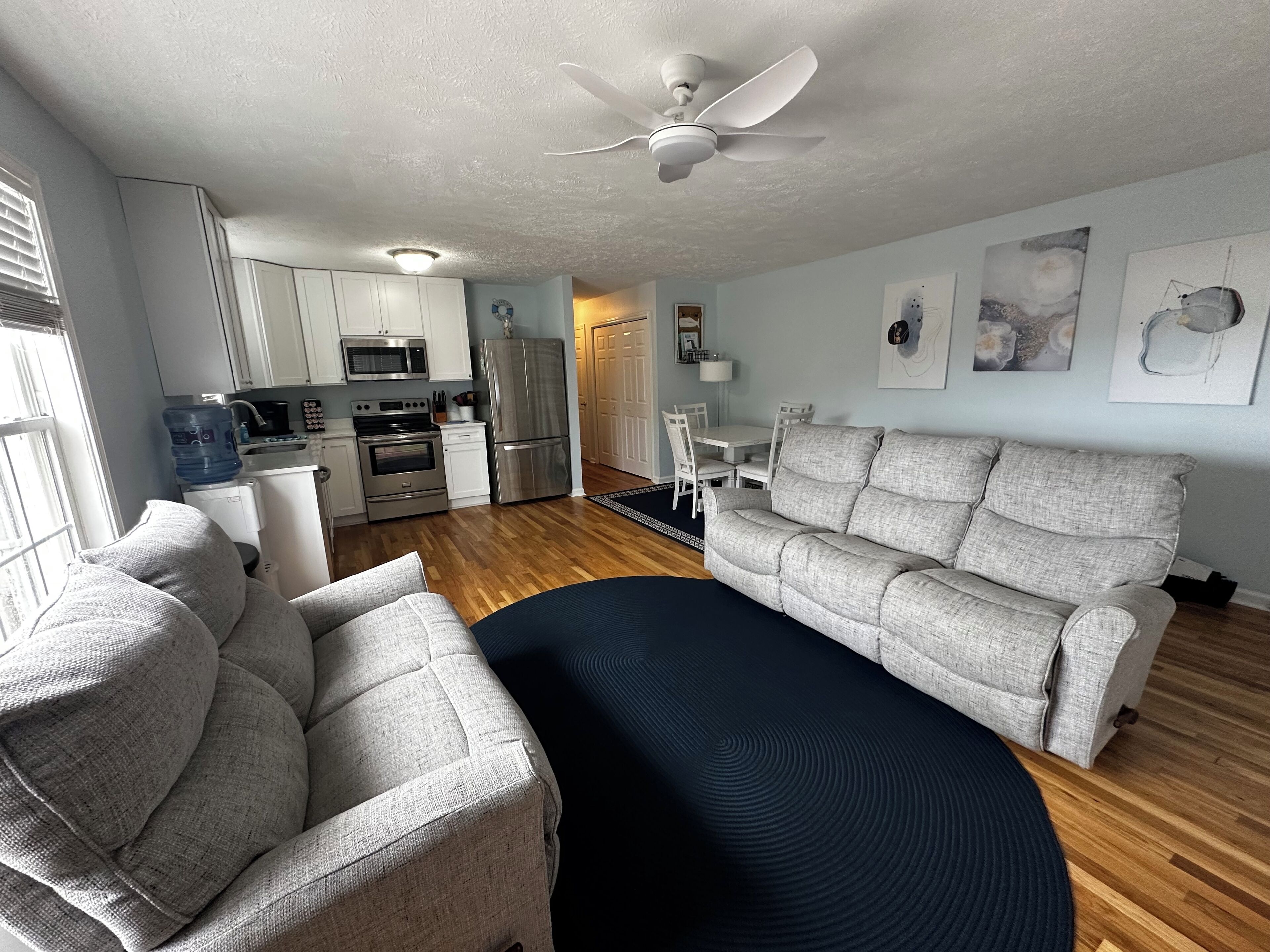 A modern living room with a grey sofa, a coffee table, and a dining area with a white table and chairs. There is a kitchen with white cabinets and stainless steel appliances.