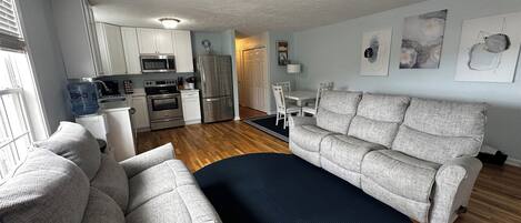 A modern living room with a grey sofa, a coffee table, and a dining area with a white table and chairs. There is a kitchen with white cabinets and stainless steel appliances.