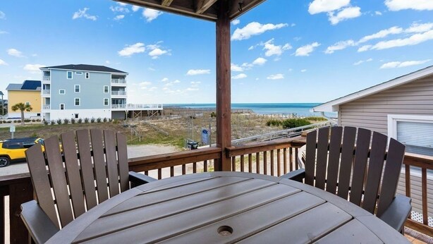 Una terraza de madera con dos sillas Adirondack y una mesa, con vistas a una playa y un edificio con varios balcones.