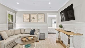 Una sala moderna con un sofá seccional color beige, una mesa de centro redonda y un mueble consola de madera. Hay tres obras de arte enmarcadas en la pared y un televisor de pantalla plana montado sobre el mueble consola.