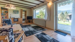 Een gezellige woonkamer met een houten plafond, een groot raam met gordijnen, een televisie, boekenkasten en een eethoek.
