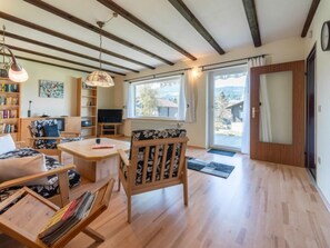 Ein geräumiges Wohnzimmer mit Holzböden, einer Couch, einem Esstisch und einem Bücherregal. Ein großes Fenster bietet einen Blick ins Freie.