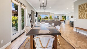 Uma ampla sala de jantar com uma mesa comprida e cadeiras, um lustre moderno e grandes janelas com vista para o exterior.