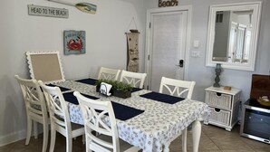 A dining area with a table set for four, surrounded by white chairs. A sign on the wall reads "HEAD TO THE BEACH."