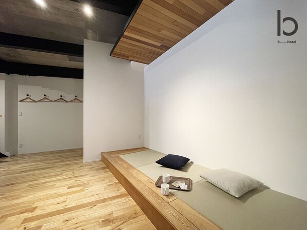 A minimalist hotel room with a wooden floor, a bed with a pillow, a tray with a cup and tissues, and a wall-mounted coat rack.