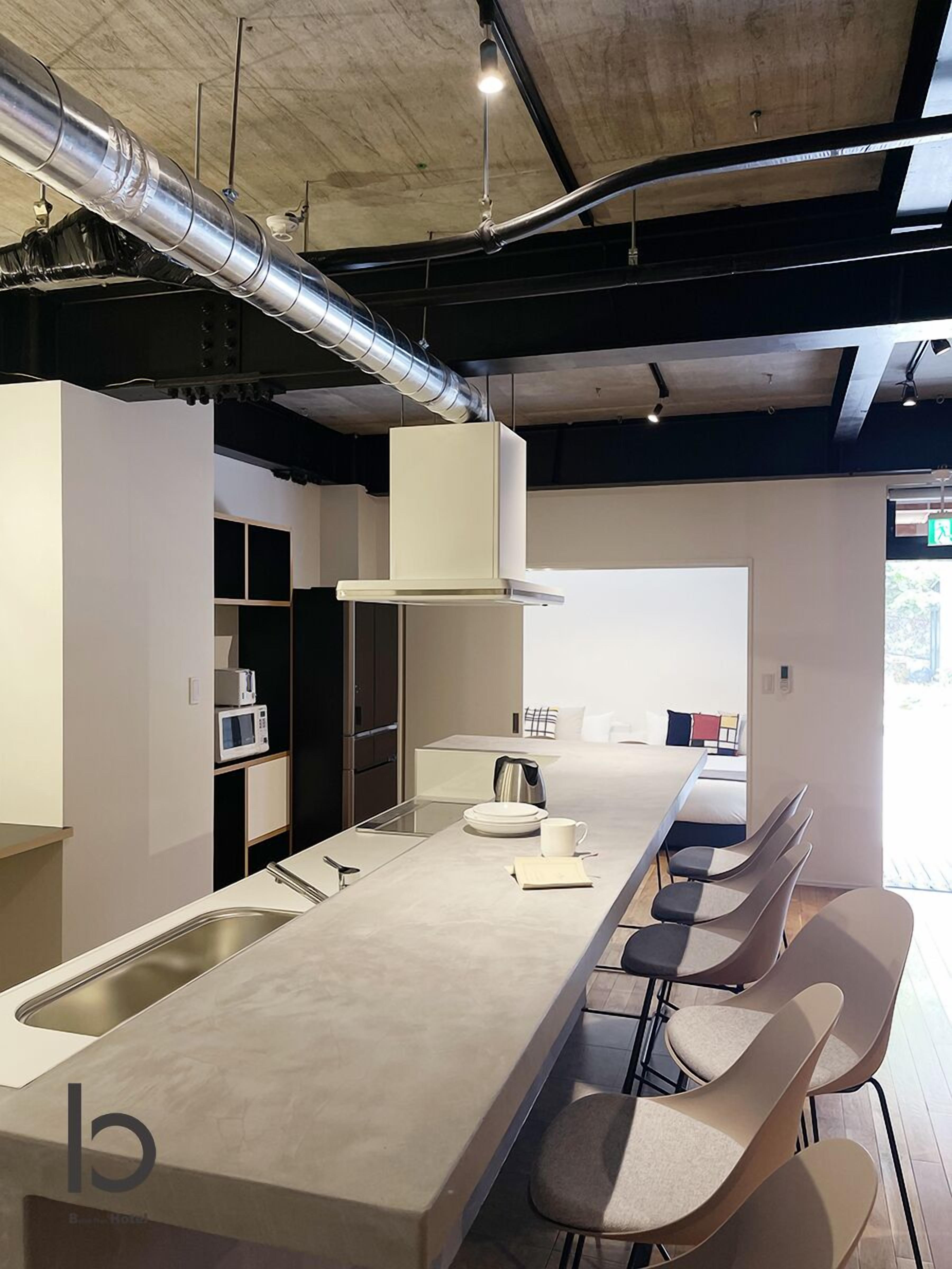 A modern kitchen with a long countertop, a built-in sink, and a stove. There are bar stools along the counter and a view of a bedroom with a bed and a window.