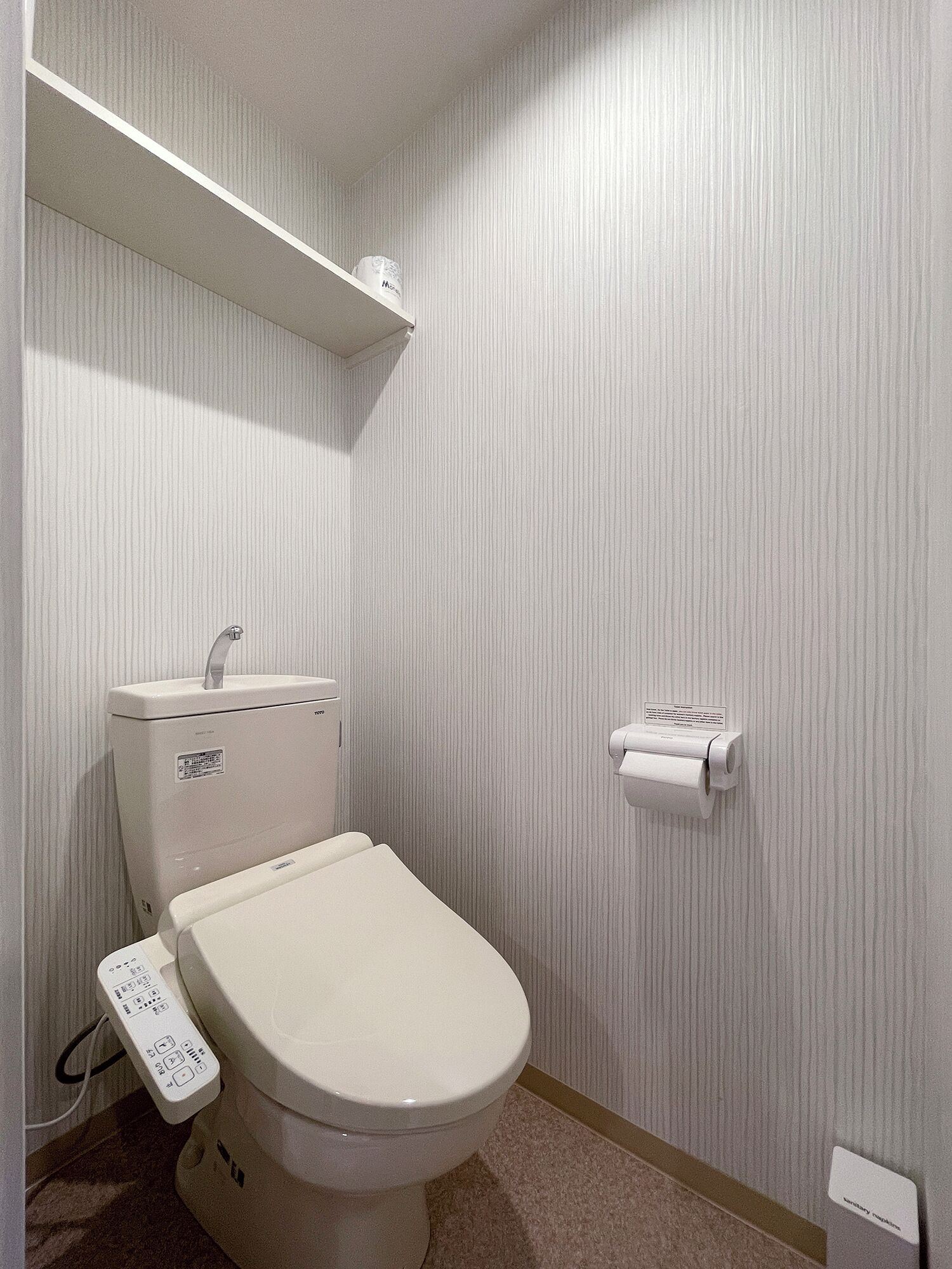 Un baño pequeño y funcional con inodoro, dispensador de papel higiénico y estante en la pared con un rollo de papel.