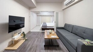 A modern hotel room with a flat-screen TV, a sofa, a wooden coffee table, and a bed with a view to the hallway. - CasaEn (Hiroshima)