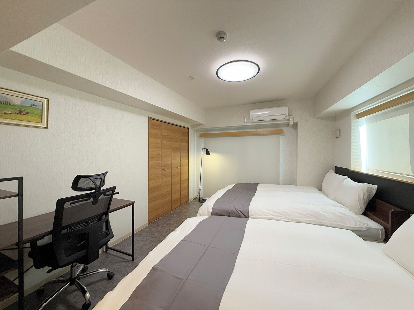 Ein Hotelzimmer mit zwei Betten, einem Schreibtisch mit Stuhl, einer Stehlampe und einer Klimaanlage.