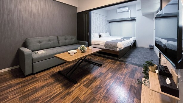 A modern hotel room with a grey sofa, a wooden coffee table, a bed with a grey headboard, and a TV on a stand. - CasaEn (Hiroshima)