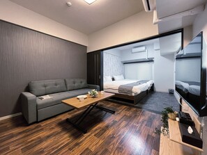 Moderni hotellihuone, jossa on harmaa sohva, puinen sohvapöytä, harmaalla päätyosalla varustettu sänky ja jalustalla oleva televisio.