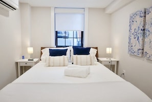 Quarto de hotel com uma cama grande, duas mesas de cabeceira com lumin árias, uma janela com persianas e um painel decorativo na parede.