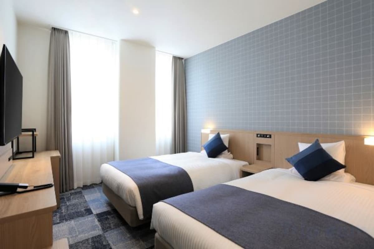 A hotel room with two beds, a desk, a television, and a window with curtains.