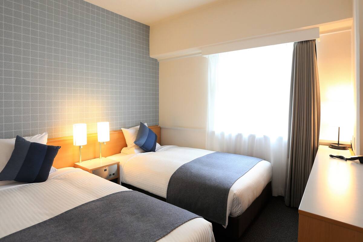 A hotel room with two beds, a nightstand with a lamp, a desk with a chair, and a window with curtains.
