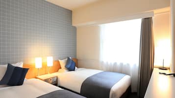 A hotel room with two beds, a nightstand with a lamp, a desk with a chair, and a window with curtains.
