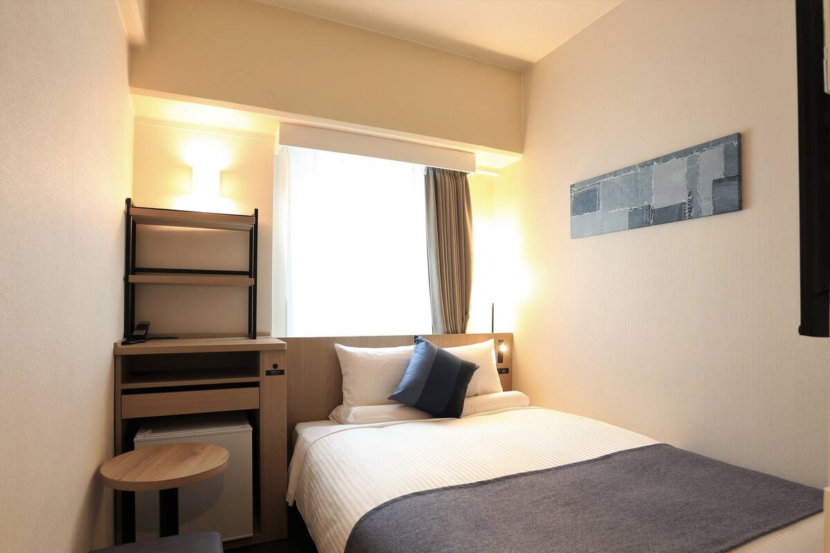A compact hotel room with a bed, a small desk, a chair, and a window with curtains.