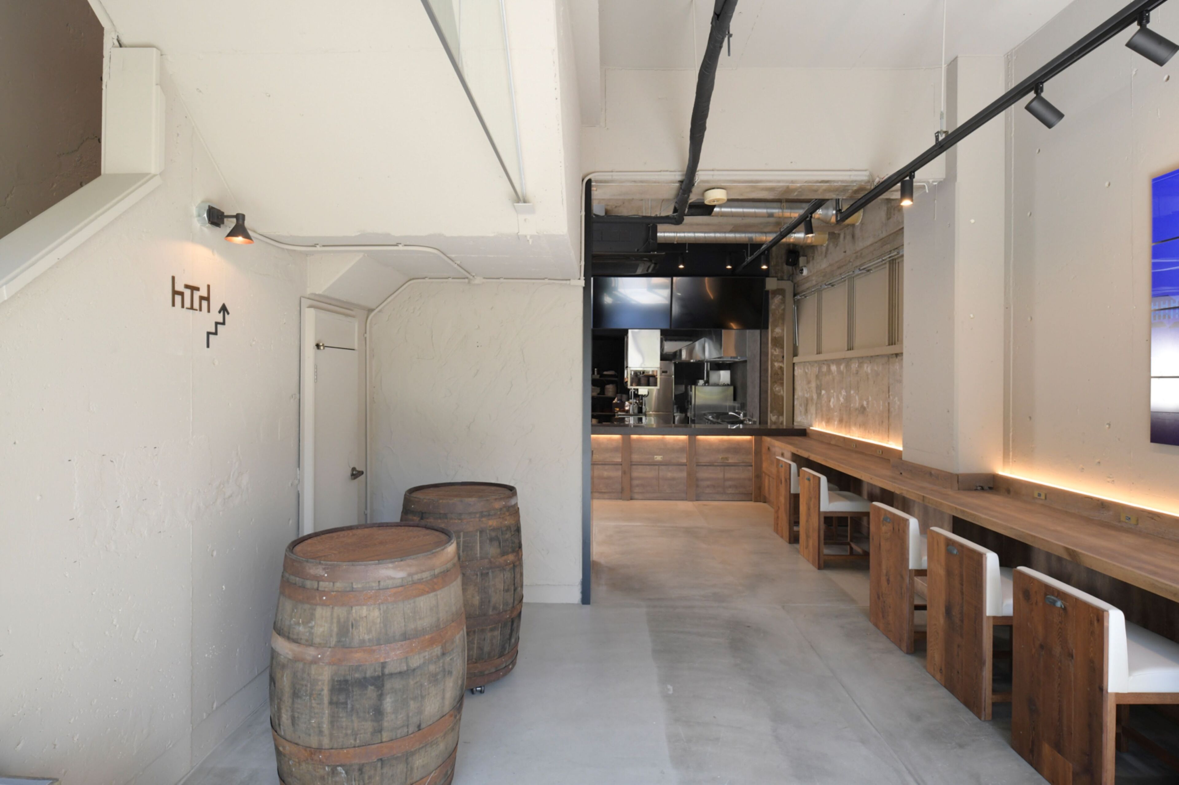 A narrow corridor with wooden barrels, a bar area with stools, and a kitchen in the background.