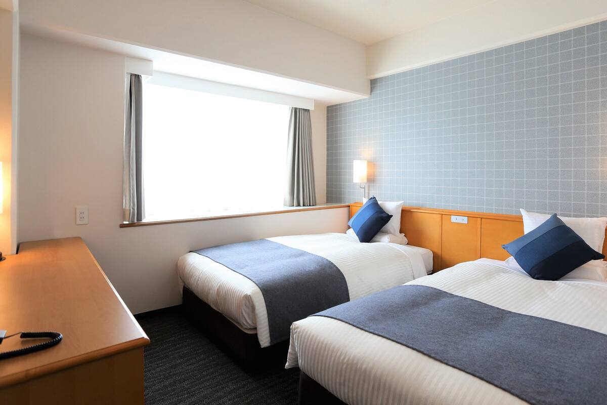 A hotel room with two beds, a desk, a window with curtains, and a wall with a grid pattern.