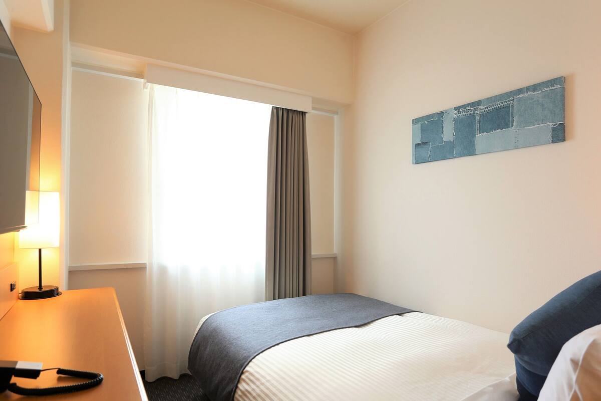 A hotel room with a bed, a desk with a lamp, a television, and a window with curtains.